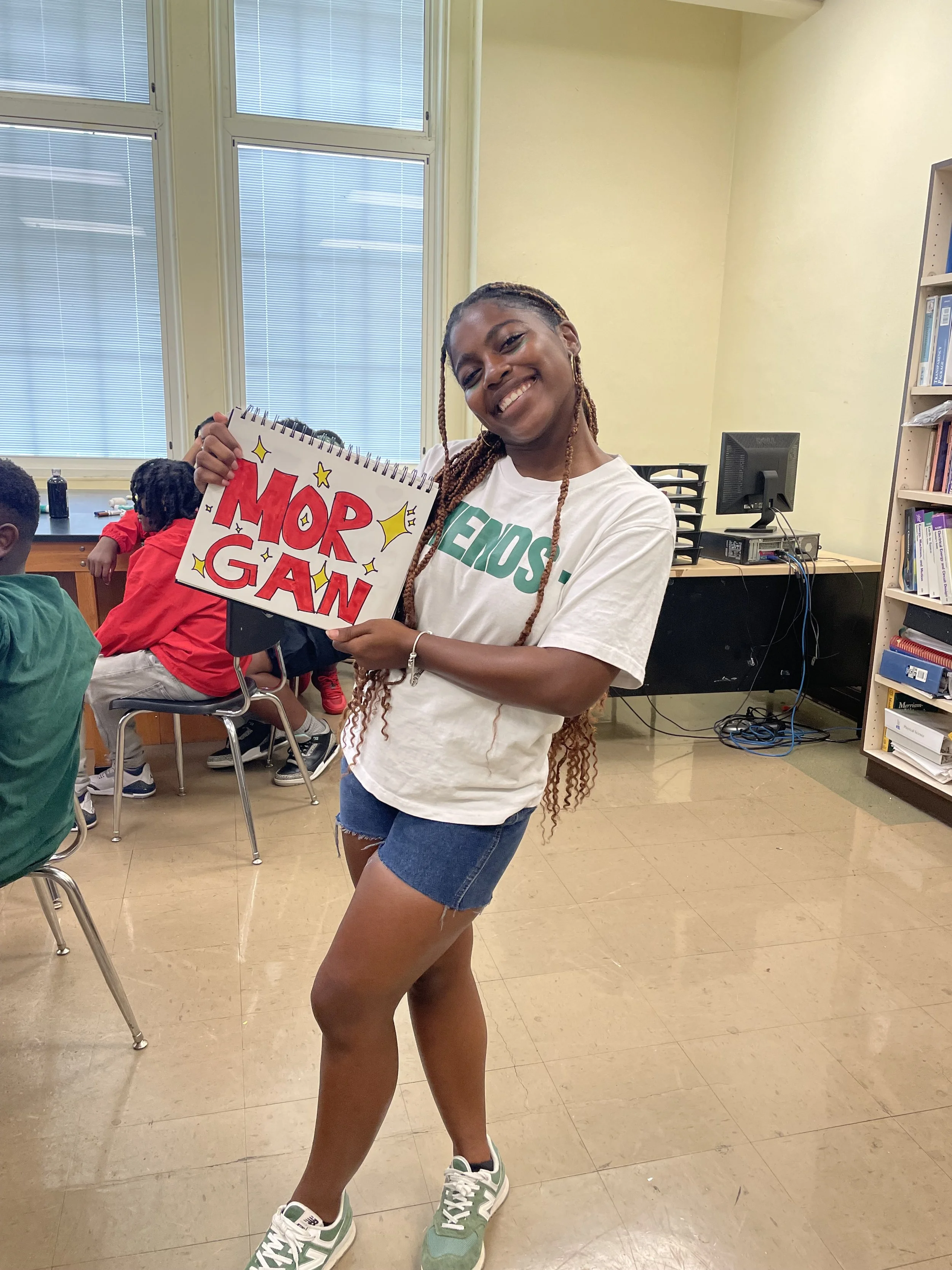 A young woman smiling and holding a sign that says 'MORE GAIN' in a classroom or office setting. She is wearing a white T-shirt, denim shorts, and green sneakers, with others and classroom furniture visible in the background.