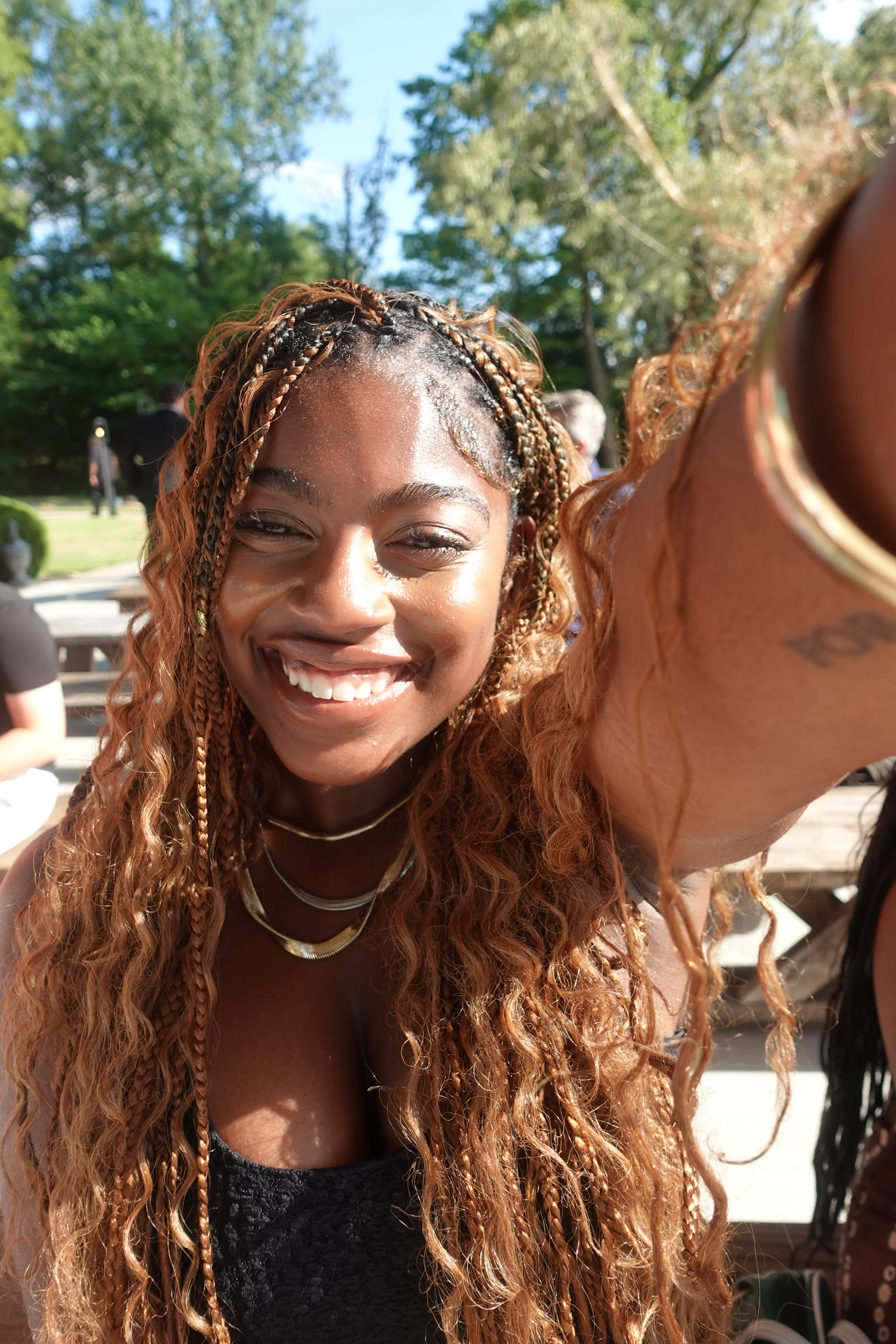 A woman with long curly hair taking a selfie outdoors, smiling broadly, wearing a black top and gold jewelry, with trees and other people in the background.