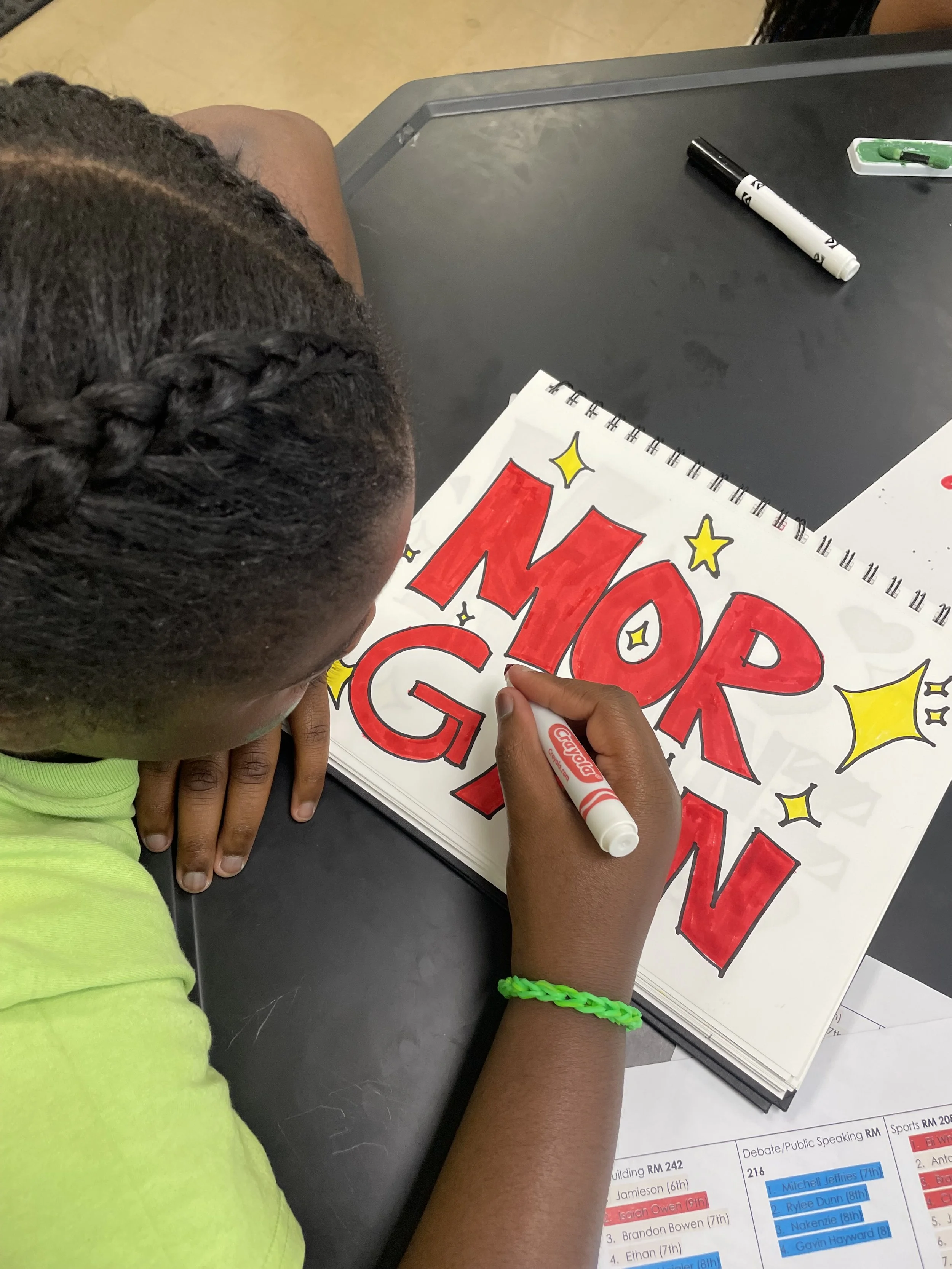 A child with braided hair and wearing a neon green shirt is coloring a sign that says 'MOP' in red with a black marker. The sign also features yellow stars around the letters. The child has a green bracelet on their wrist.