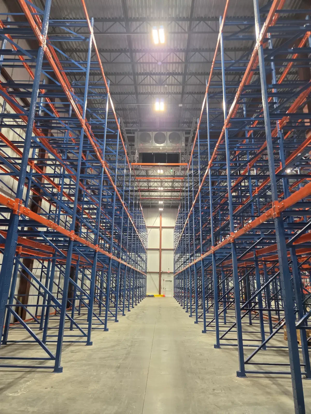 Empty warehouse with tall blue and orange metal shelving units arranged in rows, high ceiling, and a concrete floor.