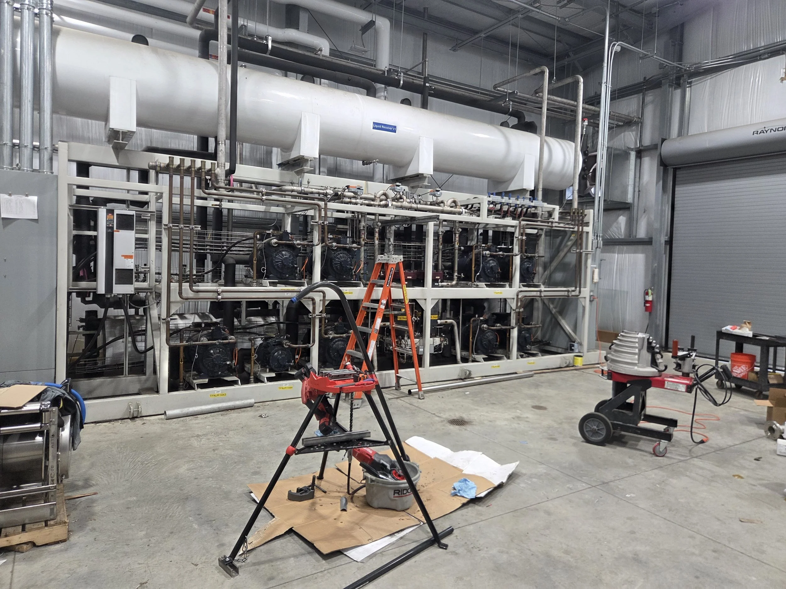 Industrial facility with large white tank labeled "Liquid Receiver V1," pipes, valves, and equipment, workshop tools, ladder, and work materials in foreground.
