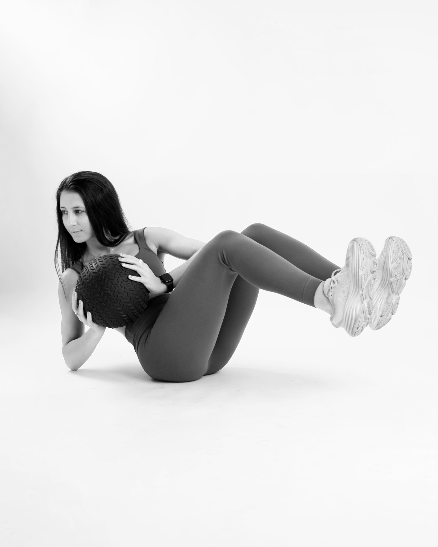 A woman in athletic clothing holding a large exercise ball, lying on her side on the floor, in a studio with a plain white background.