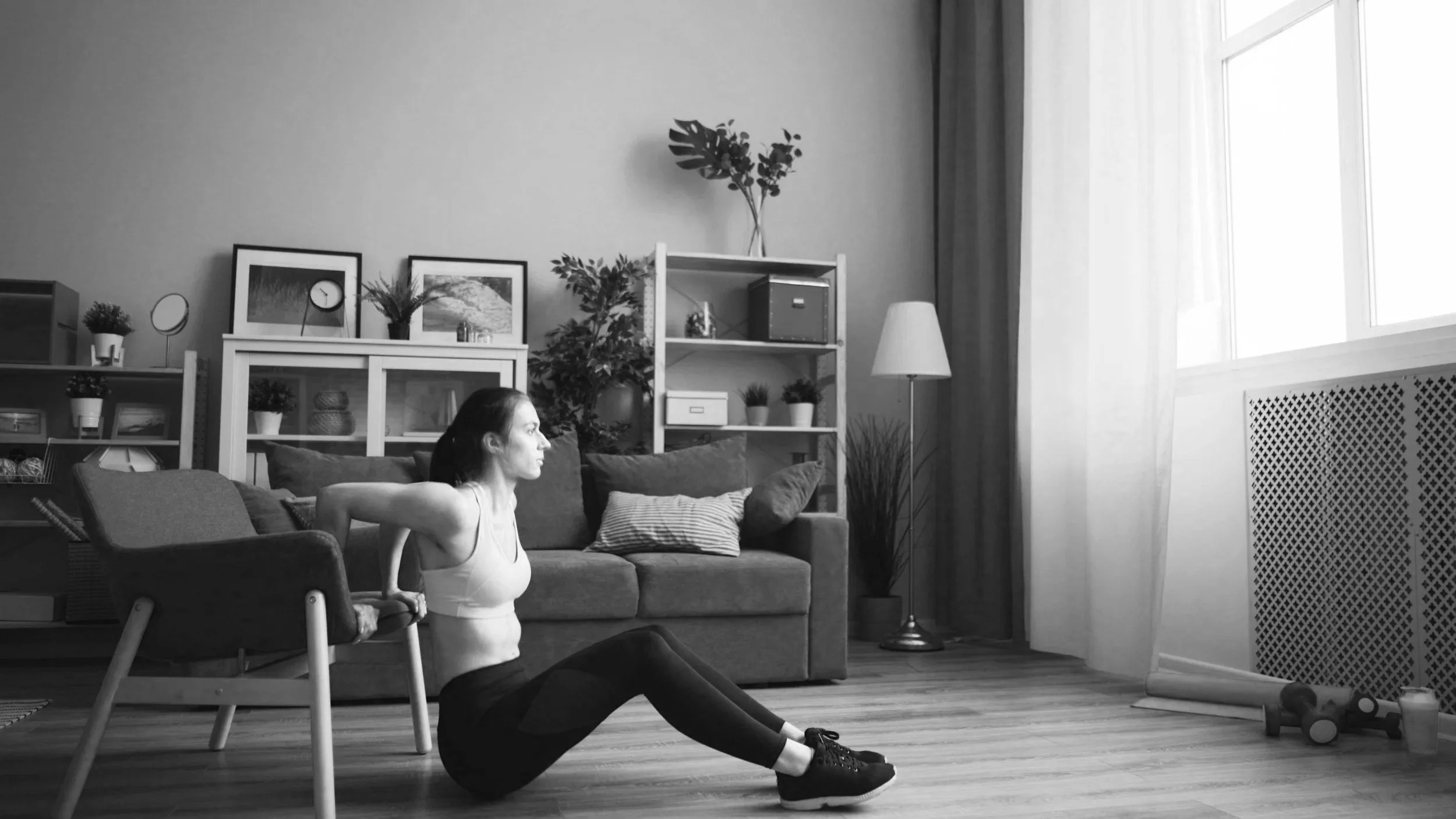 A woman exercises in a living room, performing sit-ups or crunches on the floor near a couch with cushions, in front of a shelf with decorative items, and near a window with curtains.
