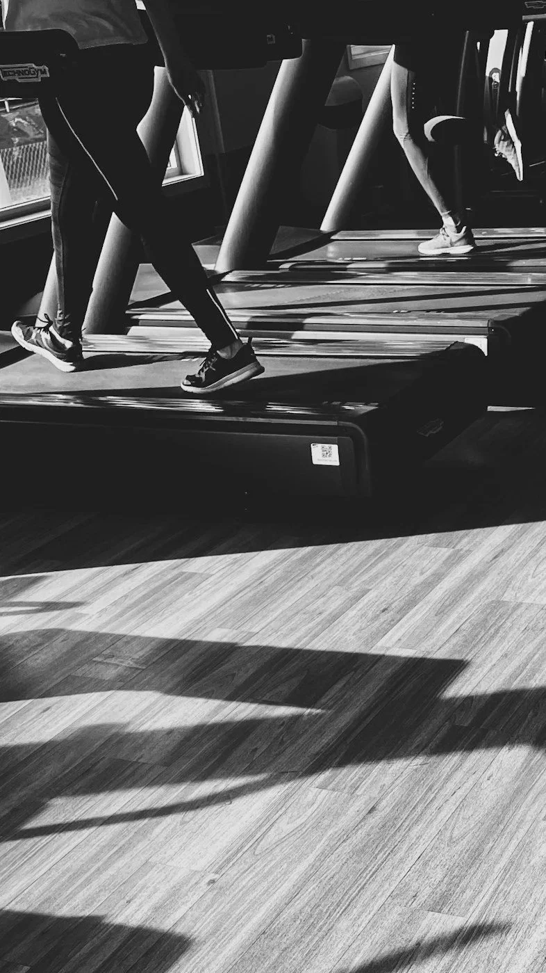People walking on treadmills in a gym, with their lower legs and feet visible, and shadows on a wooden floor.