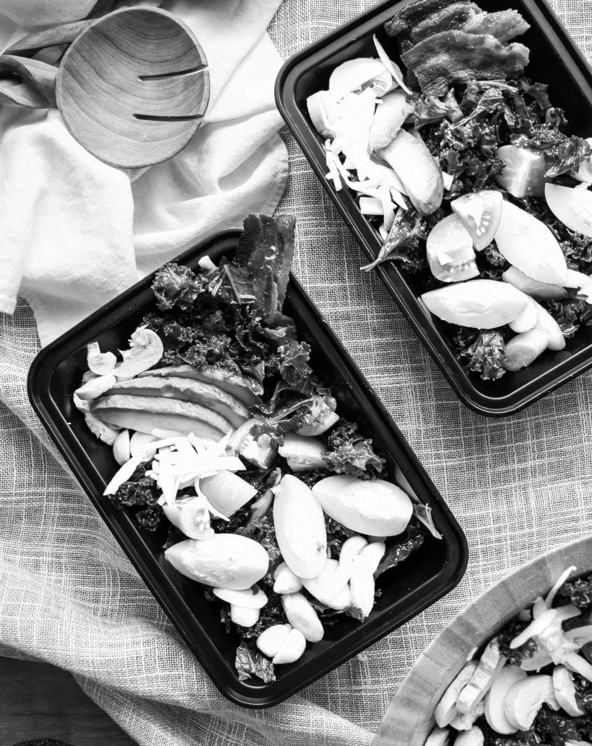 Black and white photograph of two food containers filled with sliced vegetables such as cucumbers, tomatoes, and lettuce, with additional sliced vegetables on a plate nearby. There is a textured tablecloth underneath and a wooden salad server on the side.