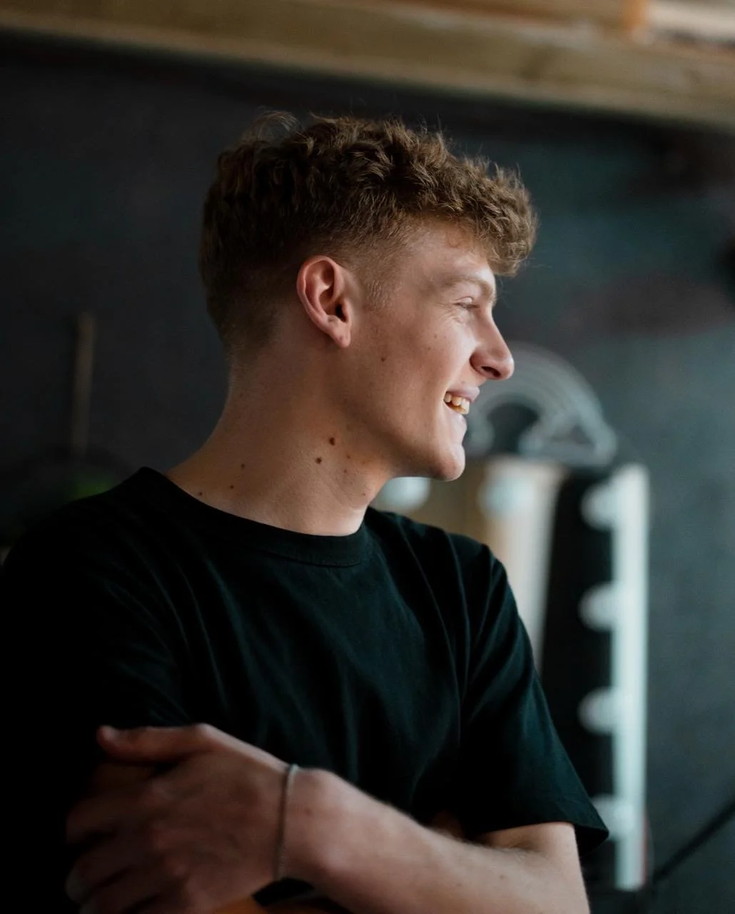 A young man with curly hair and a black t-shirt smiling and looking to his right, standing indoors with a dark background.