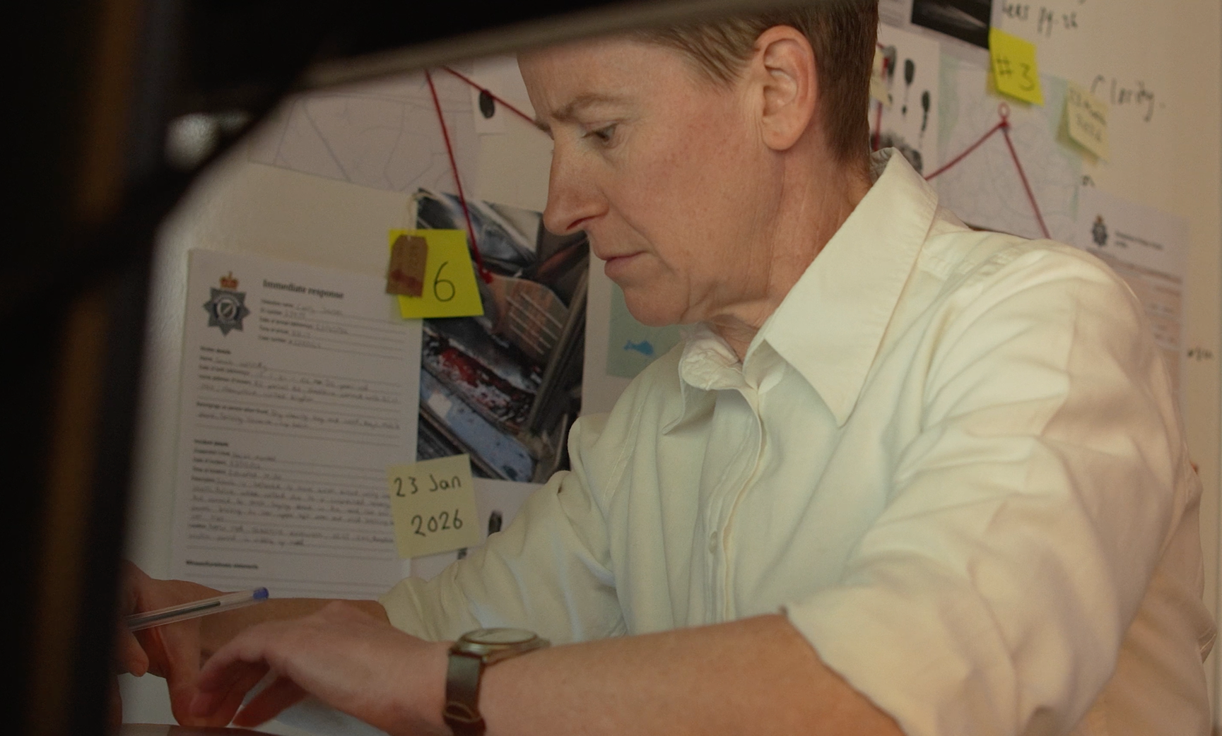 A man with short red hair wearing a white dress shirt and wristwatch, sitting at a desk and writing on a piece of paper, with a notice and photos pinned on a wall behind him. The wall also has various notes and a red string connecting some points.