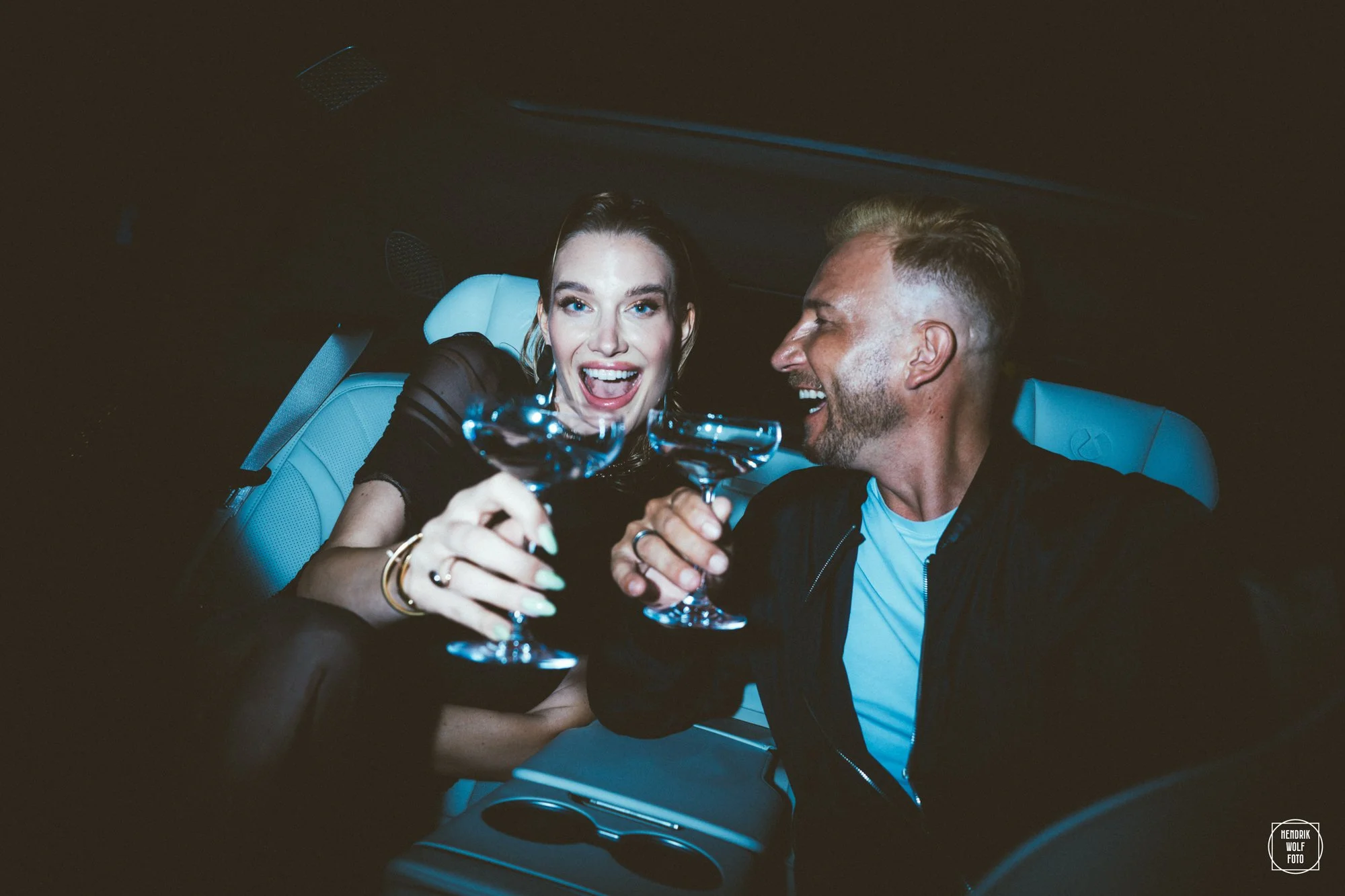 A woman and a man in a car celebrating, holding glasses of champagne and smiling at each other.