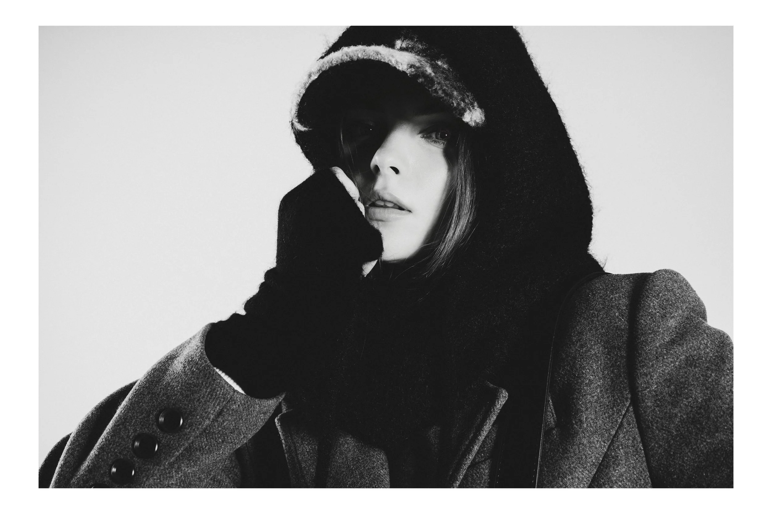 Black and white portrait of a woman wearing a wide-brimmed hat, coat, and gloves, with her face partially obscured by the hat and her hand near her face.