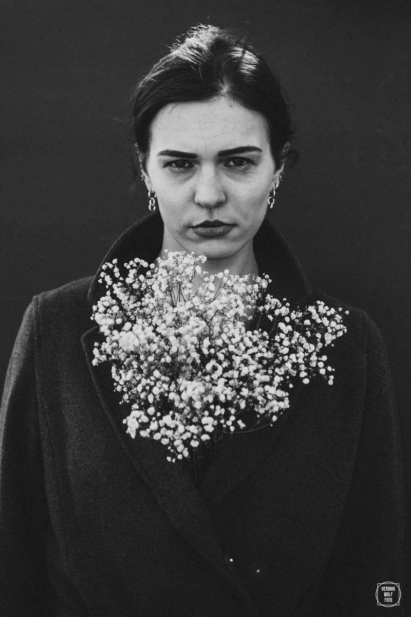 Black and white portrait of a woman with dark hair, intense gaze, and earrings, holding a bouquet of small white flowers, against a dark background.