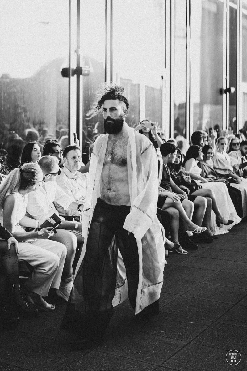 A man with a beard, long hair, and partial chest hair, wearing black pants and an open long white coat, walking among seated audience members at a fashion show, with large glass windows and reflections in the background.