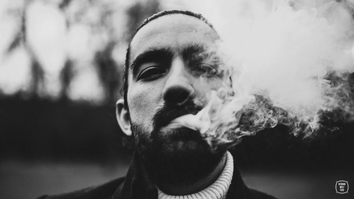 A close-up black and white portrait of a man with a beard smoking, with smoke covering part of his face.