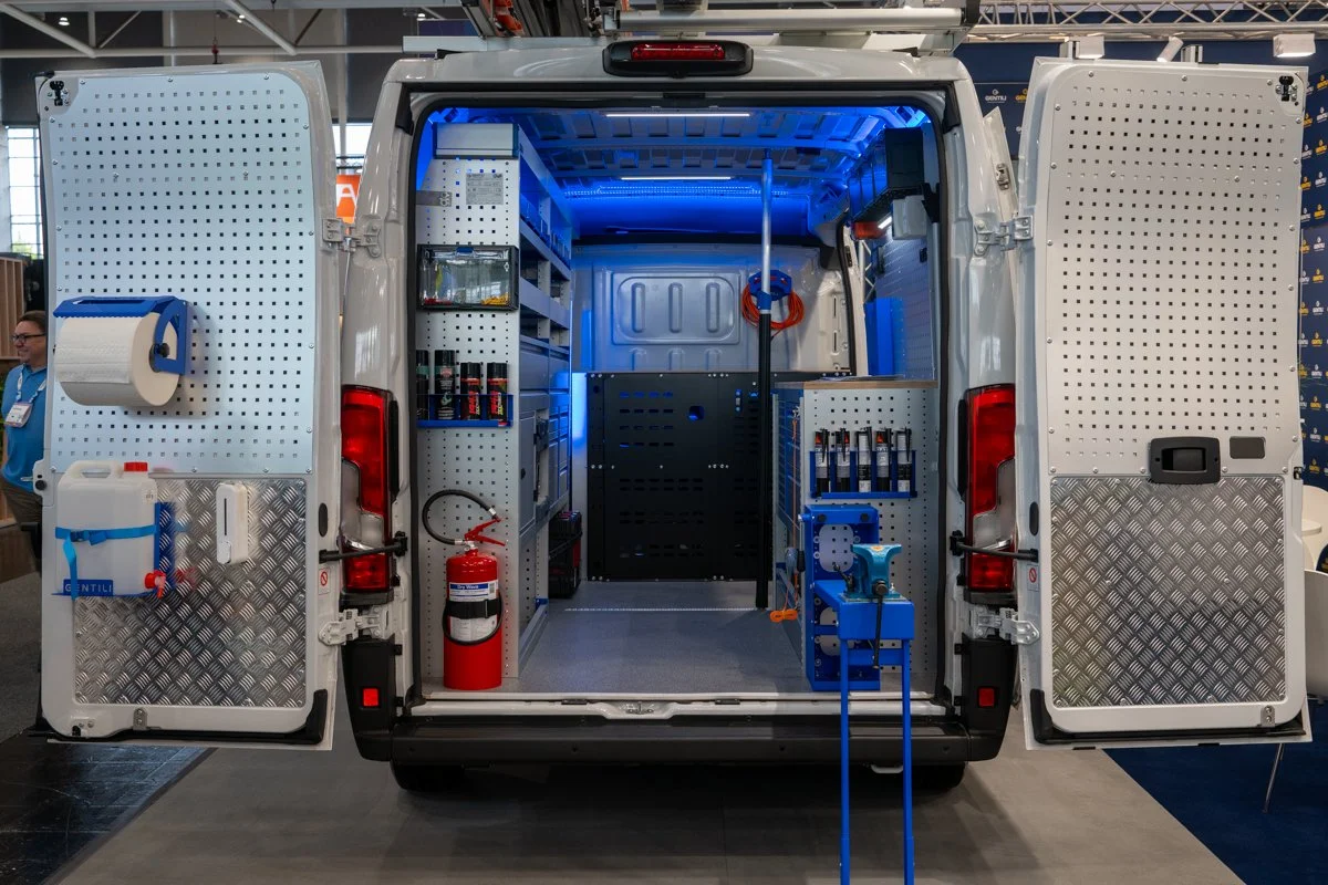 An interior view of a mobile workshop van with open doors, showing shelves with tools, fire extinguisher, paper towel dispenser, and various equipment and supplies inside.
