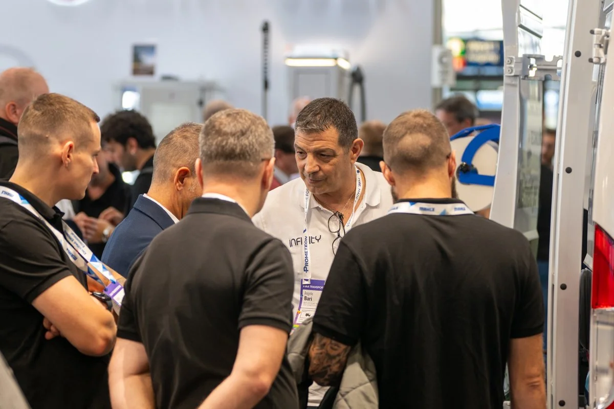 A group of men gathered around a speaker at a conference or trade show, engaging in conversation. The setting appears to be an indoor expo or convention.
