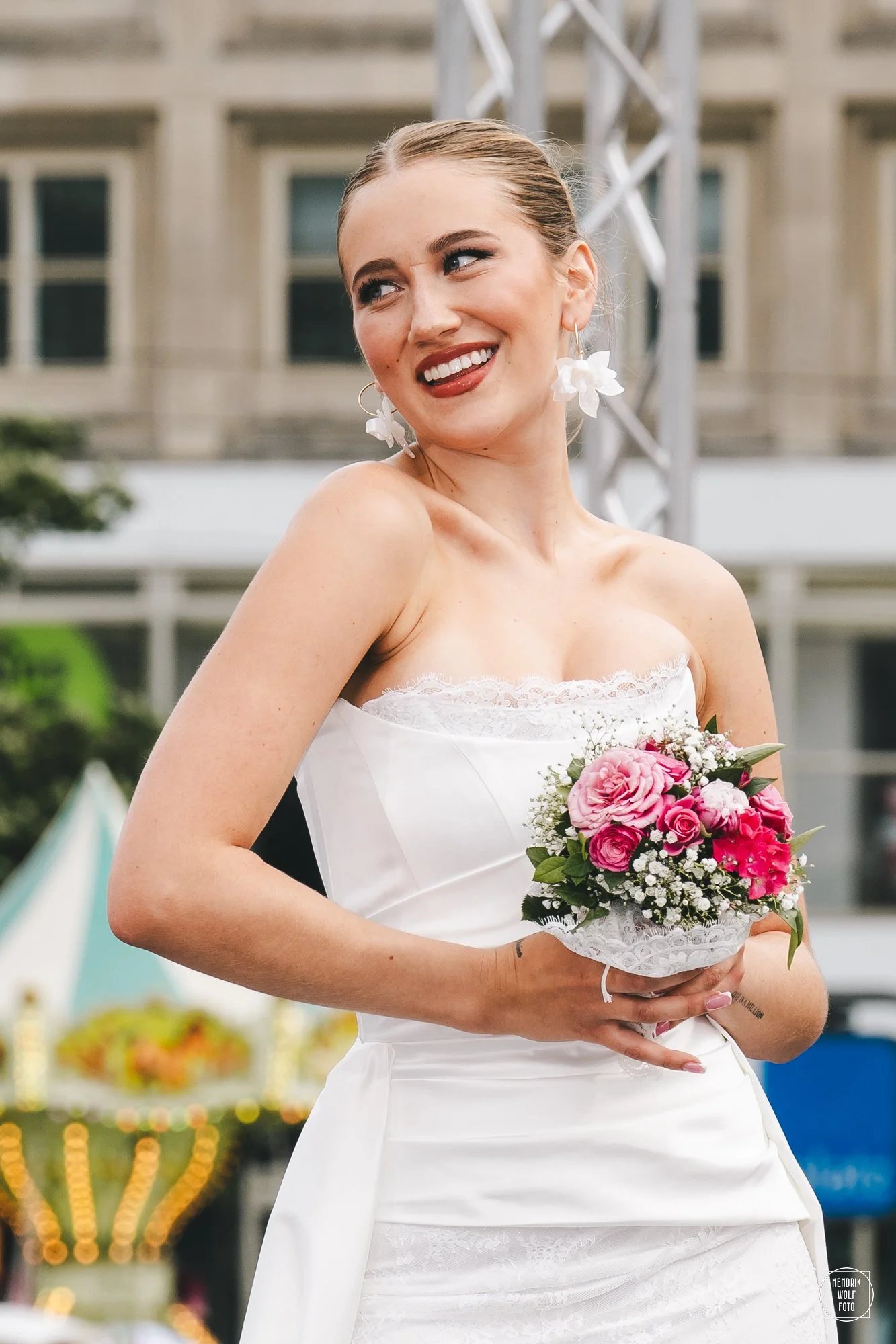 A woman in a white wedding dress holding a bouquet of pink and white flowers, smiling and posing outdoors in front of a blurred background with a amusement park or carnival scene.