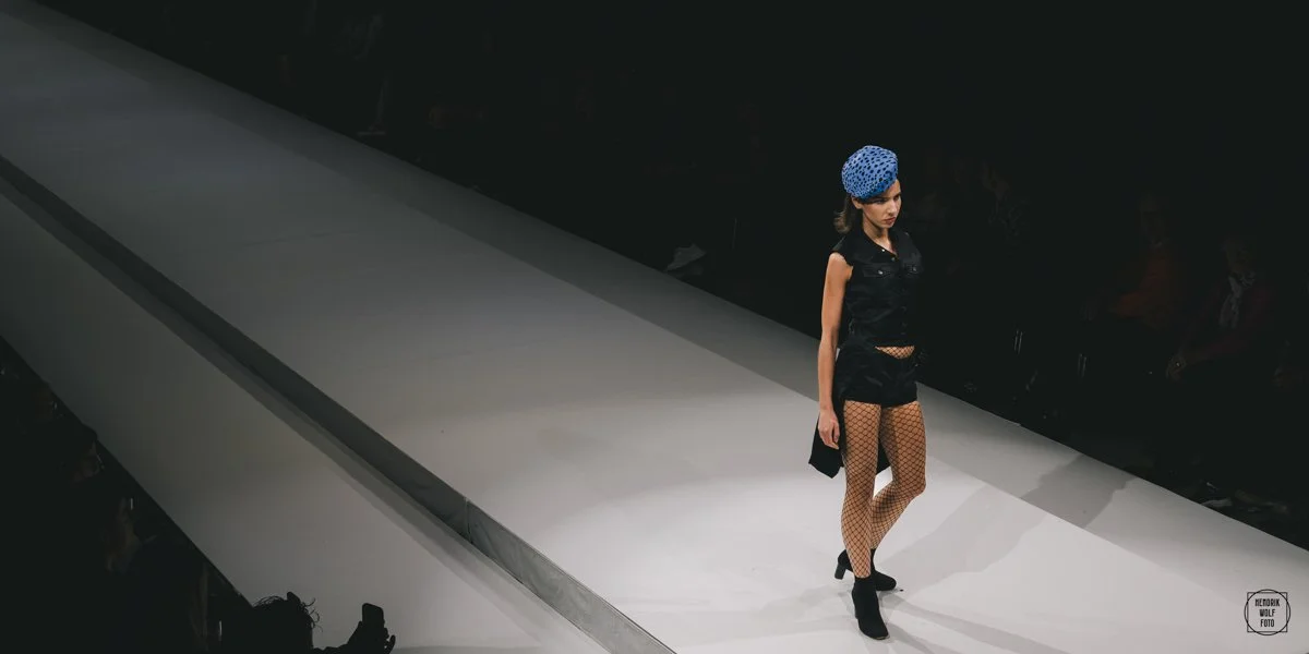 A female fashion model walks down a runway during a fashion show, wearing a black sleeveless outfit, fishnet stockings, high heels, and a blue dotted headscarf.
