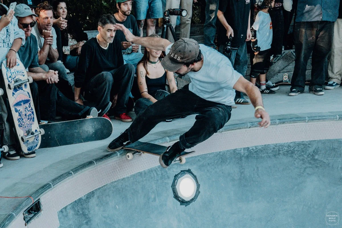 Skateboarder in white t-shirt and dark pants performing a trick on the edge of a pool, surrounded by a crowd of spectators.
