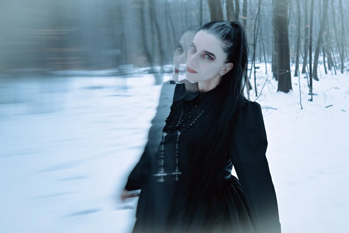 A woman with long black hair and makeup, dressed in black, standing in a snowy forest with a blurred double exposure effect.