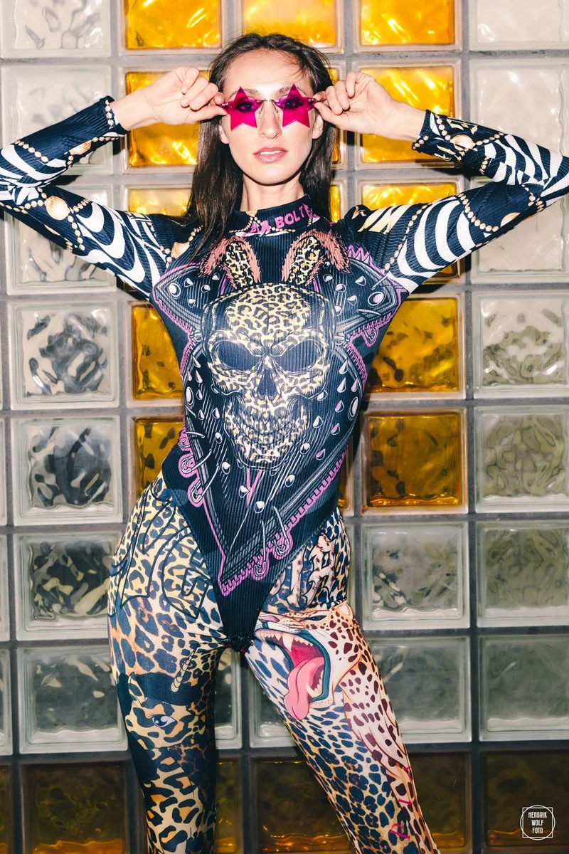 A woman wearing pink star-shaped sunglasses, a graphic long-sleeve shirt with a skull and leopard print, and matching leopard print pants with a tongue design on one leg, stands in front of a decorative glass block wall with yellow and clear glass blocks.