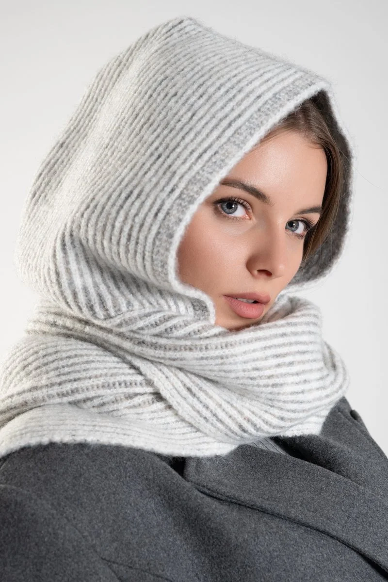 Woman wearing a gray hoodie with a hood up and wrapped in a striped scarf, looking at the camera.