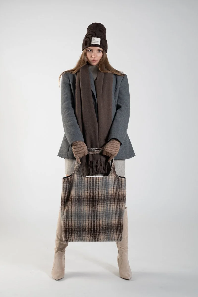 A woman wearing a brown knit beanie, gray coat, brown scarf, tan gloves, beige boots, and holding a large plaid tote bag, standing against a plain background.