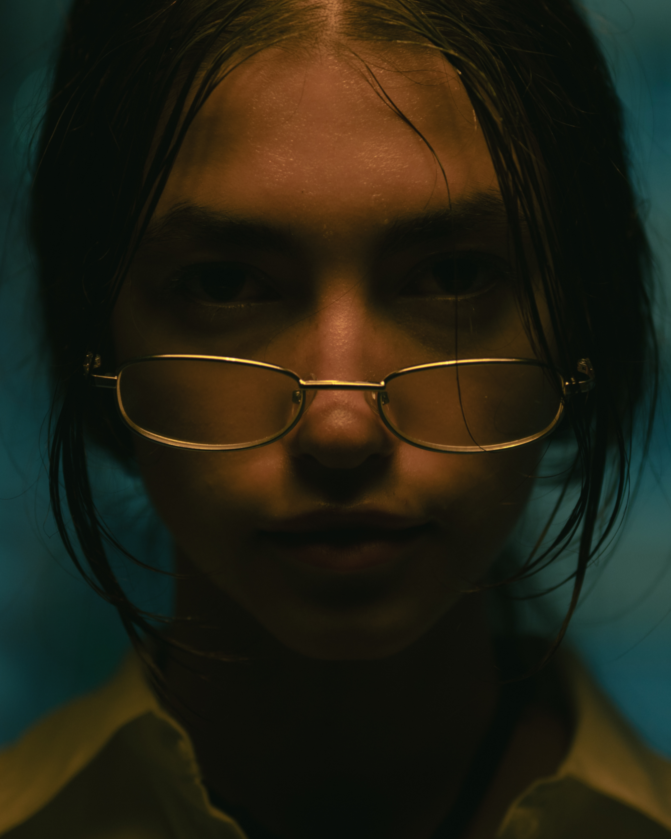 Close-up of a person's face with wet dark hair, glasses, and partially closed eyes, with a dark background.