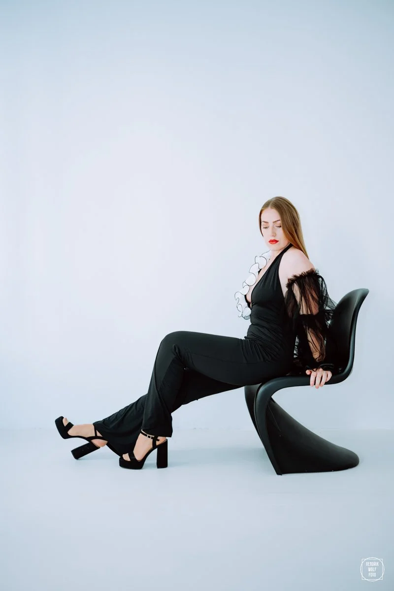 A woman dressed in black with red lipstick sitting sideways on a modern black chair in front of a plain white background.