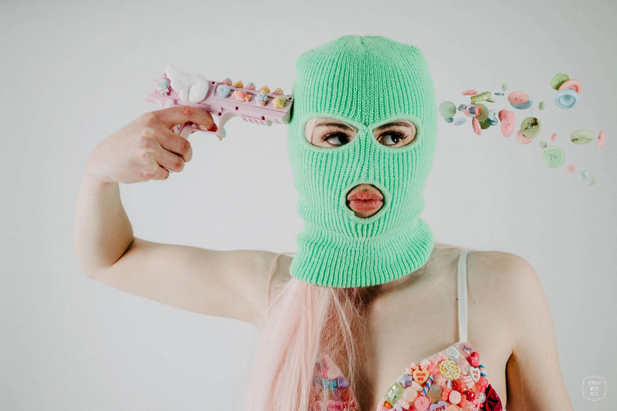 Person wearing a bright green knit balaclava with eye openings, holding a toy gun to their head, and surrounded by colorful candies and buttons. The person has pinkish hair and is wearing a dress decorated with sweets and messages.