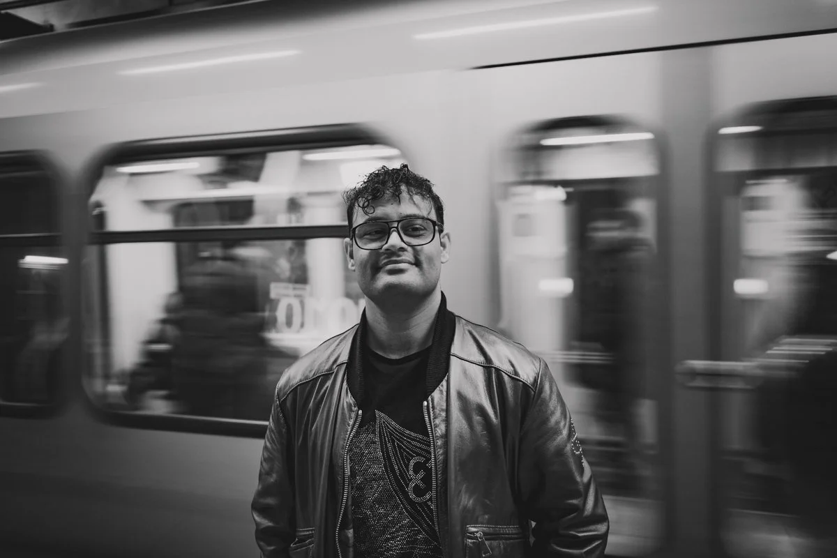 A young man wearing glasses and a leather jacket stands in front of a moving train at a subway station, captured in black and white.