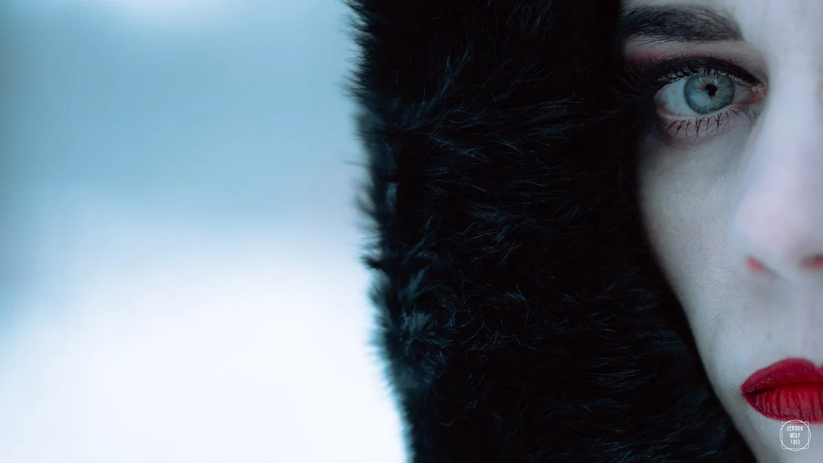 Close-up of a woman's face with one eye visible, red lipstick,  wearing a black fur-lined hood covering part of her face.