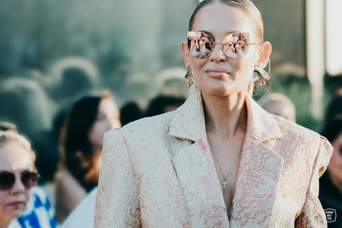 A woman with light skin wearing pink reflecting sunglasses, earrings, a cross necklace, and a light-colored blazer with a textured pattern, standing outdoors with a blurred crowd in the background.