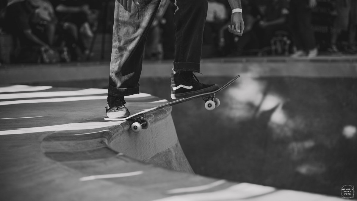 A skateboarder in black pants and sneakers is performing a trick on the edge of a skate bowl or ramp at an indoor skatepark
