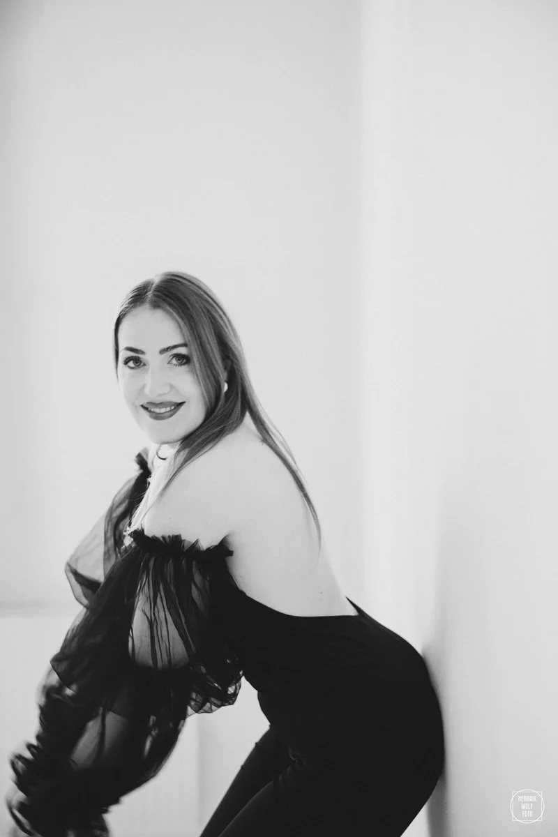 A woman with long, straight hair smiling while leaning forward against a wall in a black off-the-shoulder dress.