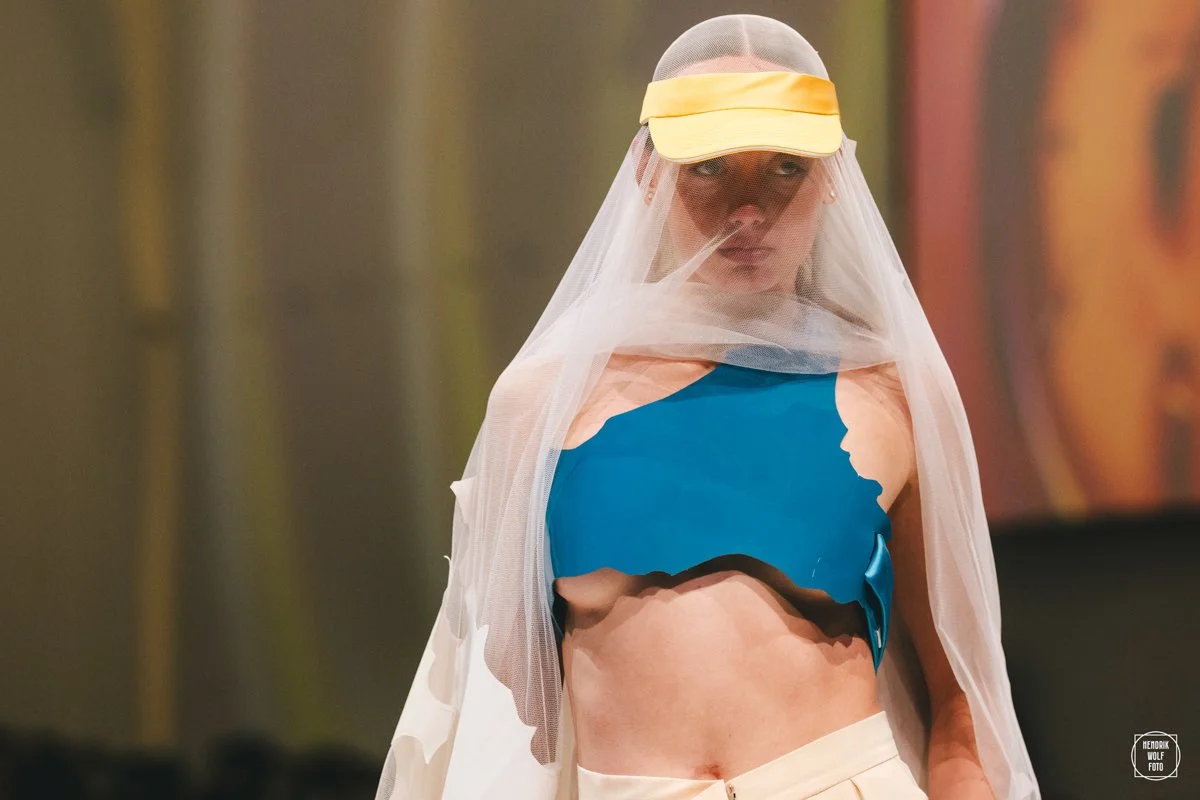 Woman wearing a yellow visor, sheer veil, torn blue top, and beige shorts, standing indoors.