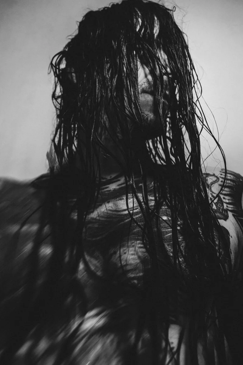 A close-up black and white photo of a person's face with long wet hair covering most of it, and a tattoo on the shoulder.