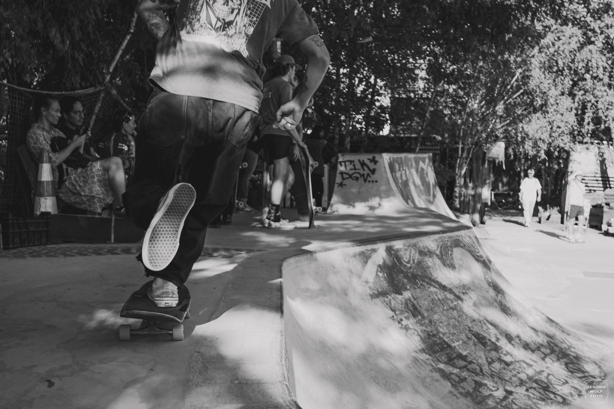 A person performing a skateboard trick at a skate park, with people watching in the background and graffiti on the ramp.