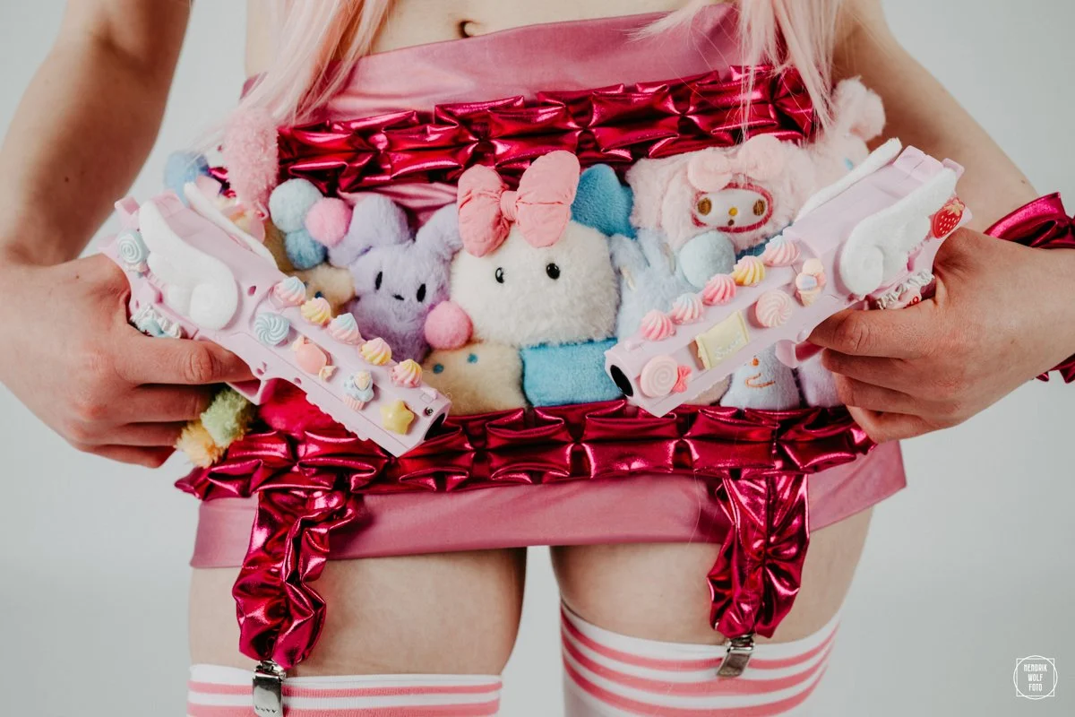 Person holding a pink box filled with colorful plush toys and decorative candy-themed items, dressed in pink and white striped stockings and a pink shiny outfit.