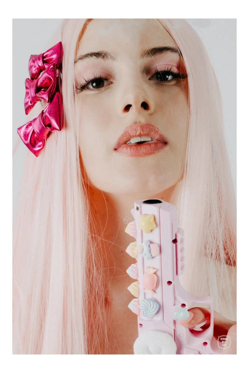 A young woman with long, straight pastel pink hair wears pink bows in her hair, has pink lipstick, and holds a pink toy gun decorated with pastel gummy candies, with a neutral background.