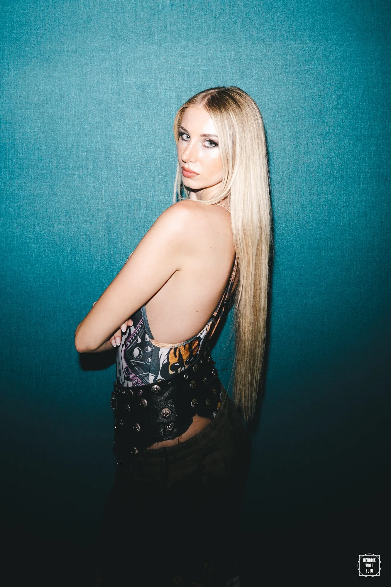 A young woman with long blonde hair standing sideways against a teal wall, looking at the camera with a serious expression, wearing a colorful graphic tank top and dark shorts with metal eyelets.