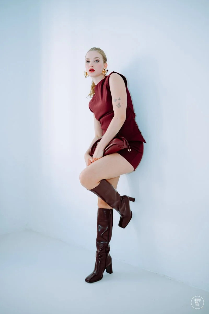 A woman in a burgundy outfit with thigh-high boots, earrings, and makeup, posing against a plain light background.