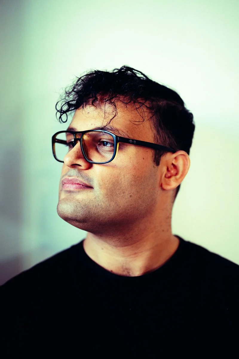 A man with short dark hair, wearing black-rimmed glasses, faces slightly to the left against a neutral background.