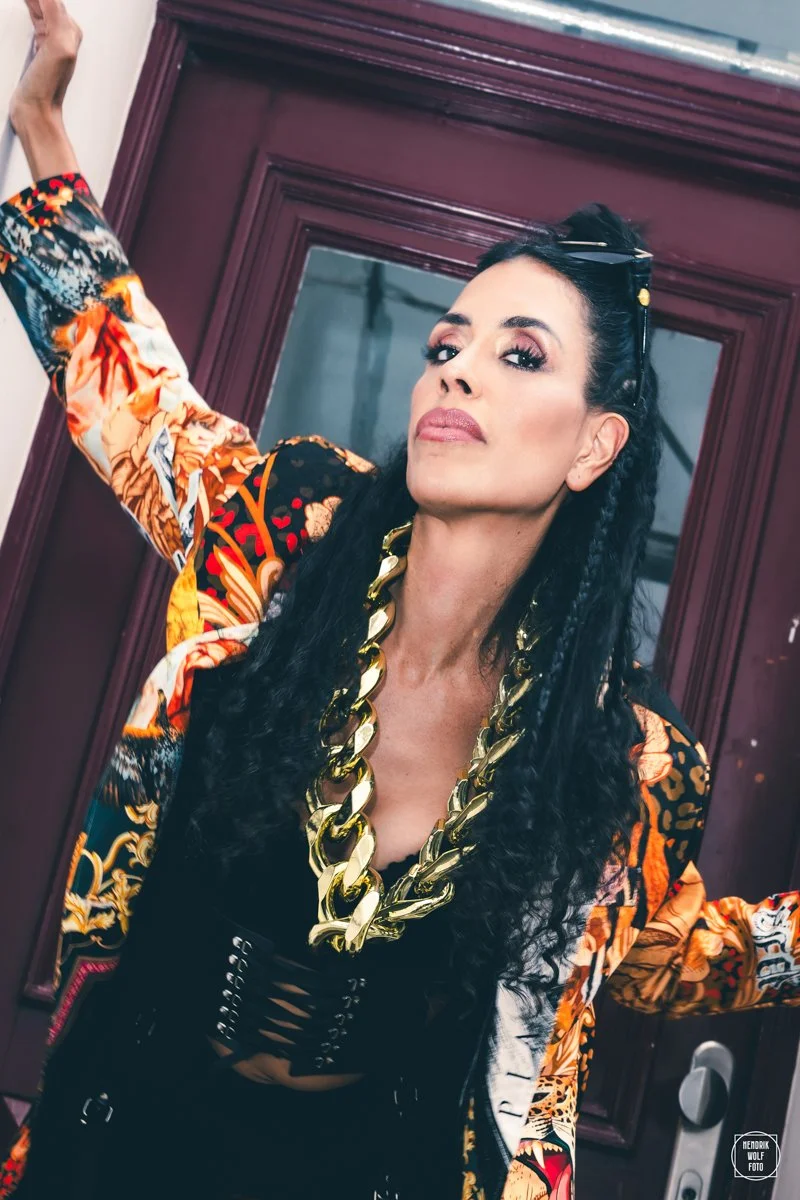 A woman with long dark curly hair, wearing heavy makeup, a large gold chain necklace, and a colorful, patterned jacket, standing in front of a wooden door.