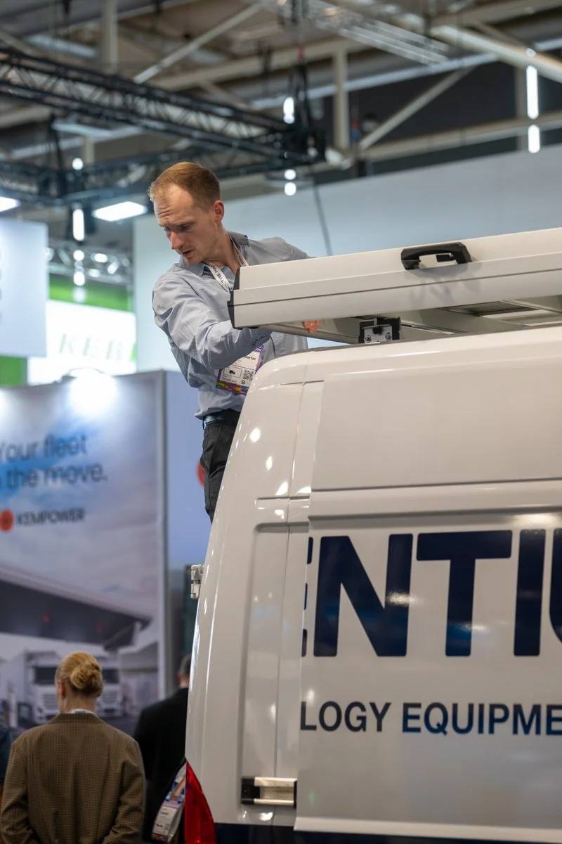 A man examining equipment in the back of a white van at an indoor trade show or conference.