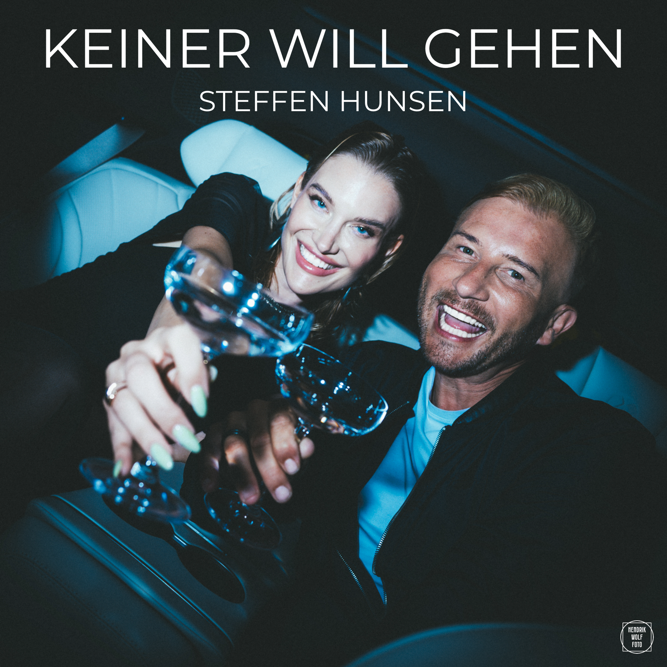 Close-up of two smiling individuals, a woman and a man, taking a selfie inside a car. The woman has blonde hair and blue eyes, and the man has short blonde hair and a beard. They are holding a champagne glass and appear joyful.