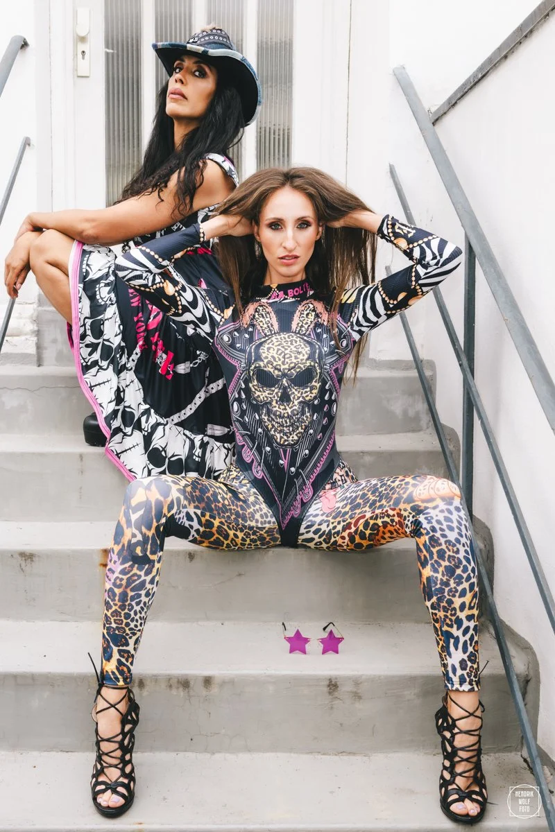 Two women with long hair posing on concrete stairs, one sitting and leaning back with legs apart, the other perched behind her. Both wear bold, colorful, animal print outfits and accessories, including a hat and sunglasses.