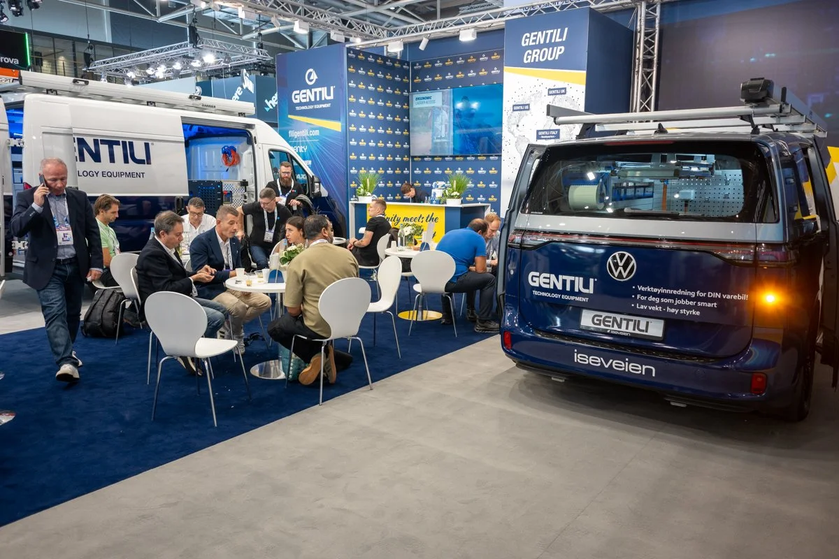 At a trade show booth, several people are seated around tables with a backdrop displaying 'GENTILI GROUP' and 'GENTIL' branding. A Volkswagen van with the 'GENTILI' logo and additional text is parked nearby, and there is a white service vehicle with 