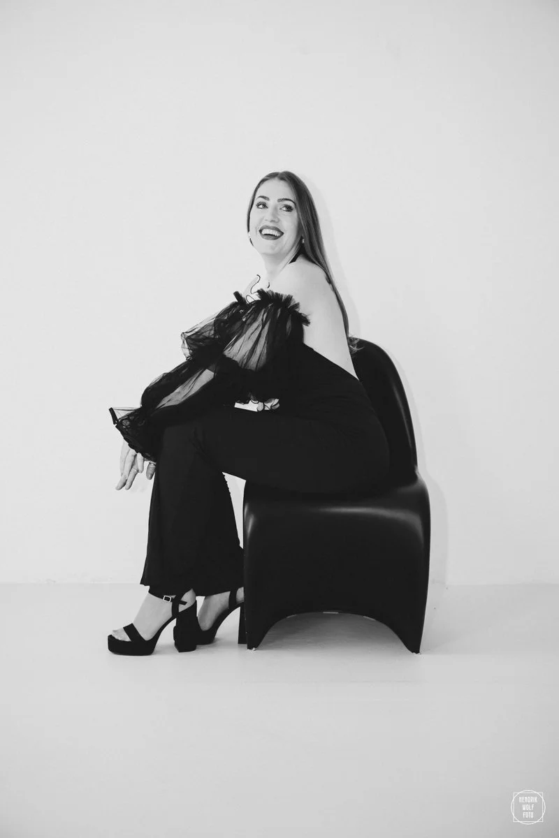 A woman sitting on a modern black chair, wearing a black dress with sheer puffed sleeves, smiling and looking at the camera, against a plain white wall.