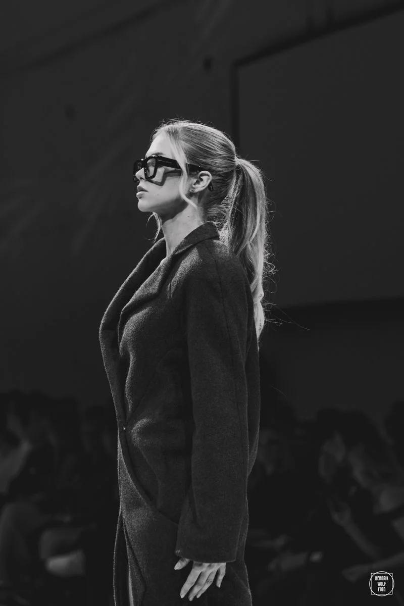 A woman with long hair tied back, wearing glasses and a dark coat, walking on a runway during a fashion show.
