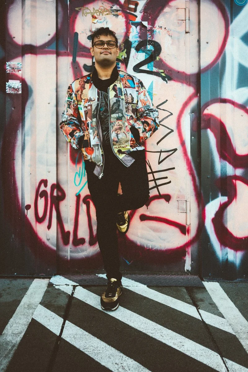 A young man standing in front of a graffiti-covered wall, wearing glasses, a colorful jacket with magazine covers, black pants, and sneakers, with his hands in his pockets and one foot crossed over the other.