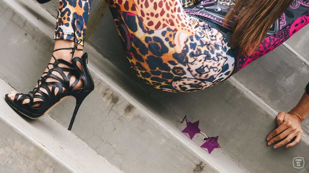 A woman wearing leopard print high heels, colorful patterned leggings, and a multicolor patterned jacket, sitting on concrete stairs with purple star-shaped sunglasses on the step.